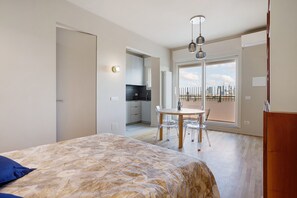 Une chambre avec un lit, une table à manger avec des chaises, un coin cuisine et une grande fenêtre donnant sur la ville.