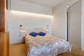 Un dormitorio con cama, mesita de noche, repisa en la pared y una puerta.