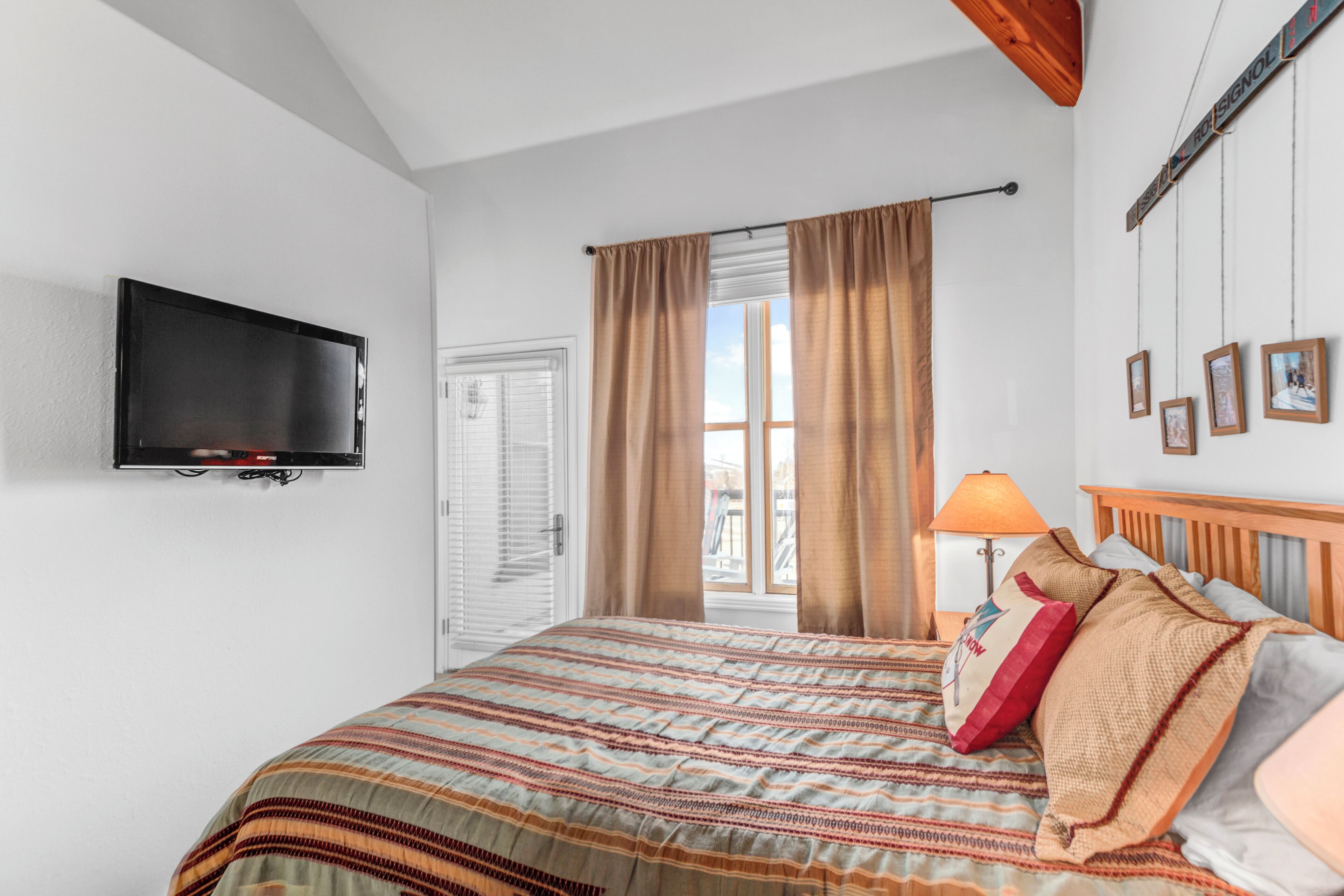 A bedroom with a bed, a television mounted on the wall, a window with curtains, and a lamp.