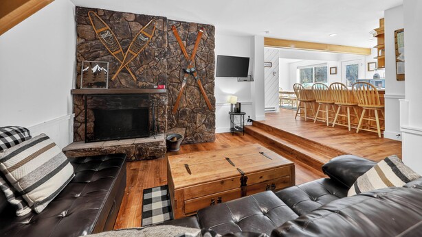 A modern living room with a stone fireplace, leather sofa, wooden dining table, and a television mounted on the wall. - Family Ski Loft at Kristi – Walk to Chair 8, Game Room, Views (Brian Head)