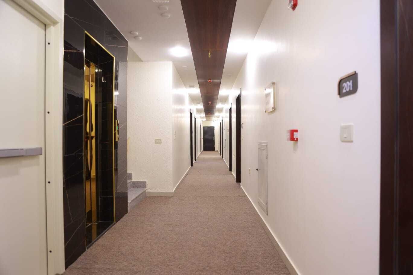 A long, narrow hallway with doors on both sides, carpeted floor, and a ceiling with recessed lighting.