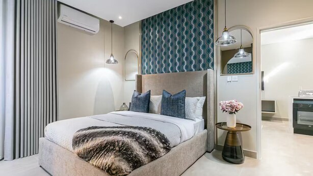 Un dormitorio moderno con una cama grande, un cabecero, una mesita de noche con un florero y vistas a la cocina.