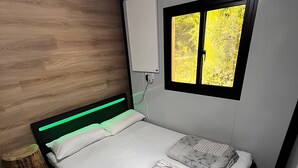 A bed with a green headboard, a bedside table, a window with a view of trees, and a wall-mounted light switch. - Tiny House 200 with Private Terrace, Garden, and Wi-Fi (Zadelsdorf)