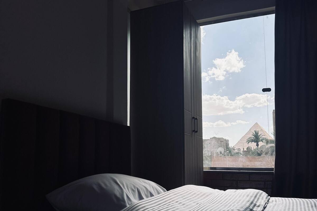 A hotel room with a bed, a window with curtains, and a view of pyramids.