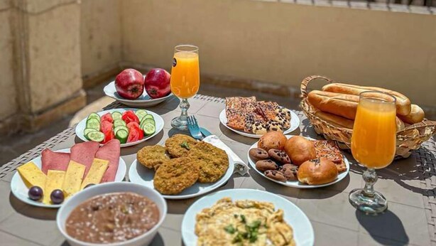 A table set with various breakfast items and a glass of orange juice on a balcony overlooking buildings and a pyramid. - The Pyramid Vault View INN (Giza)