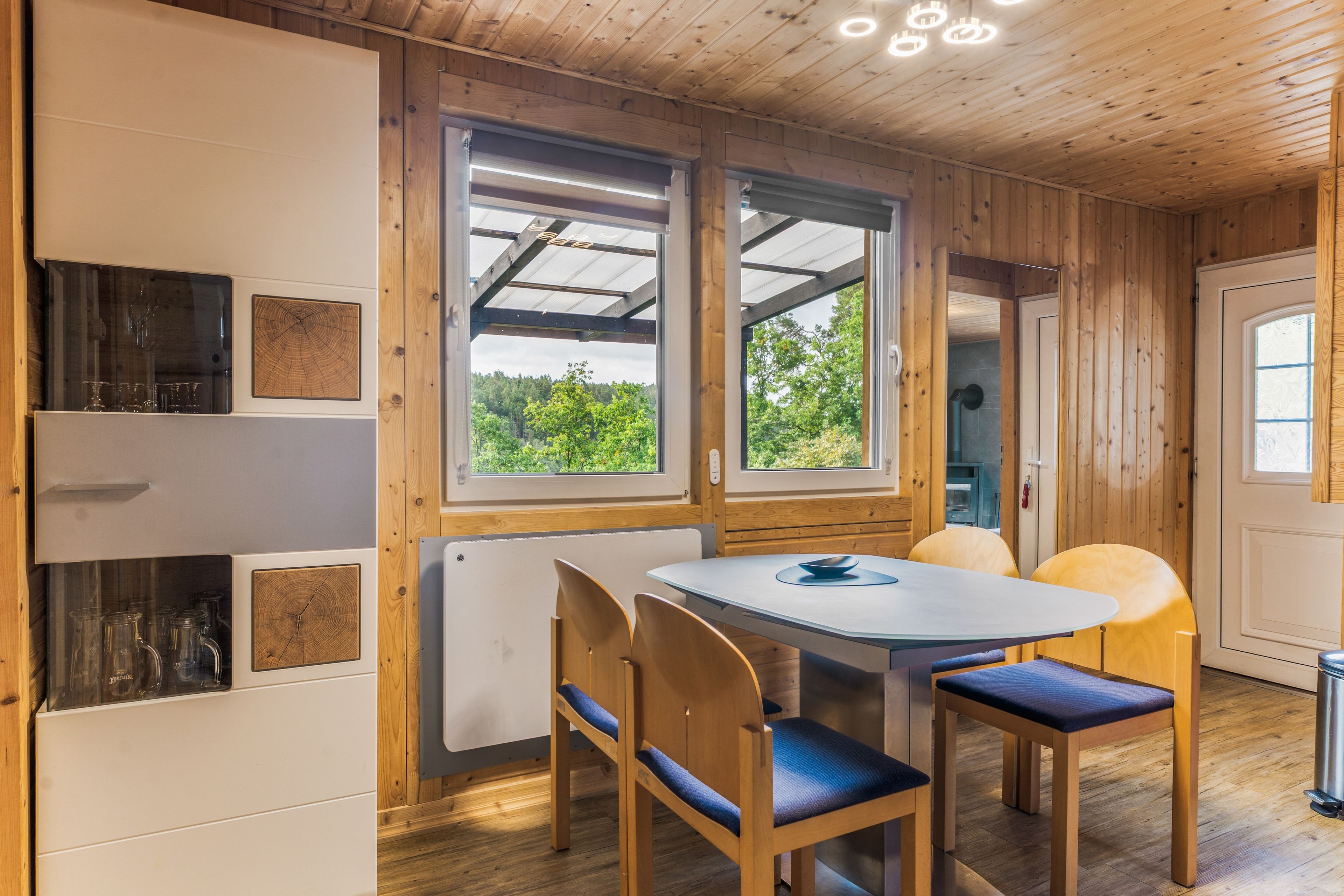 Een houten interieur met een ronde tafel, stoelen en uitzicht op groen.