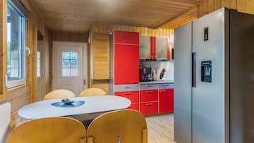 A modern kitchen with wooden walls, a stainless steel refrigerator, red cabinets, and a dining area with a white table and wooden chairs.