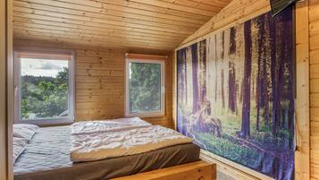 A wooden cabin bedroom with a bed, two windows, and a forest mural on the wall.