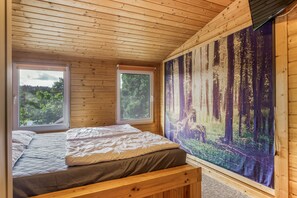 Ein Schlafzimmer einer Holzhütte mit einem Bett, zwei Fenstern und einer Waldmalerei an der Wand.