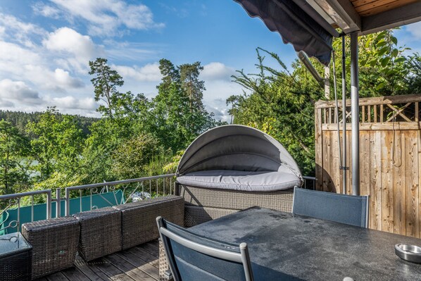 Un espace extérieur aménagé, abrité et doté de meubles en osier et d’une table, offrant une vue sur un paysage boisé.