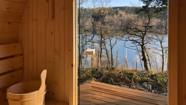 Una sauna de madera con un balde y un banco de madera, y vistas a un lago con árboles y una terraza de madera.