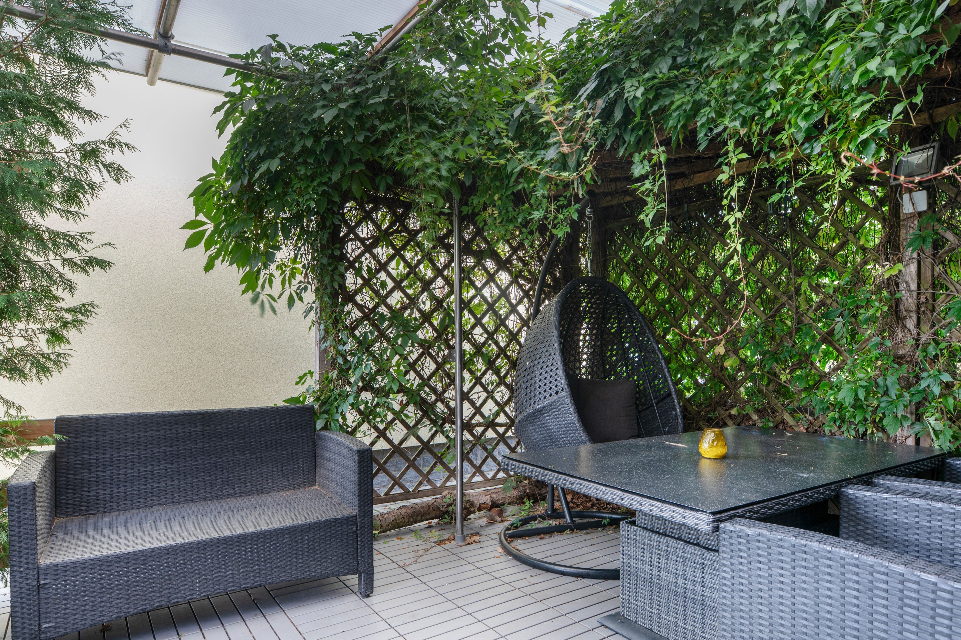 An outdoor patio area with wicker furniture and a table, surrounded by dense foliage and a lattice structure.