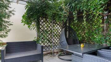 An outdoor patio area with wicker furniture and a table, surrounded by dense foliage and a lattice structure.
