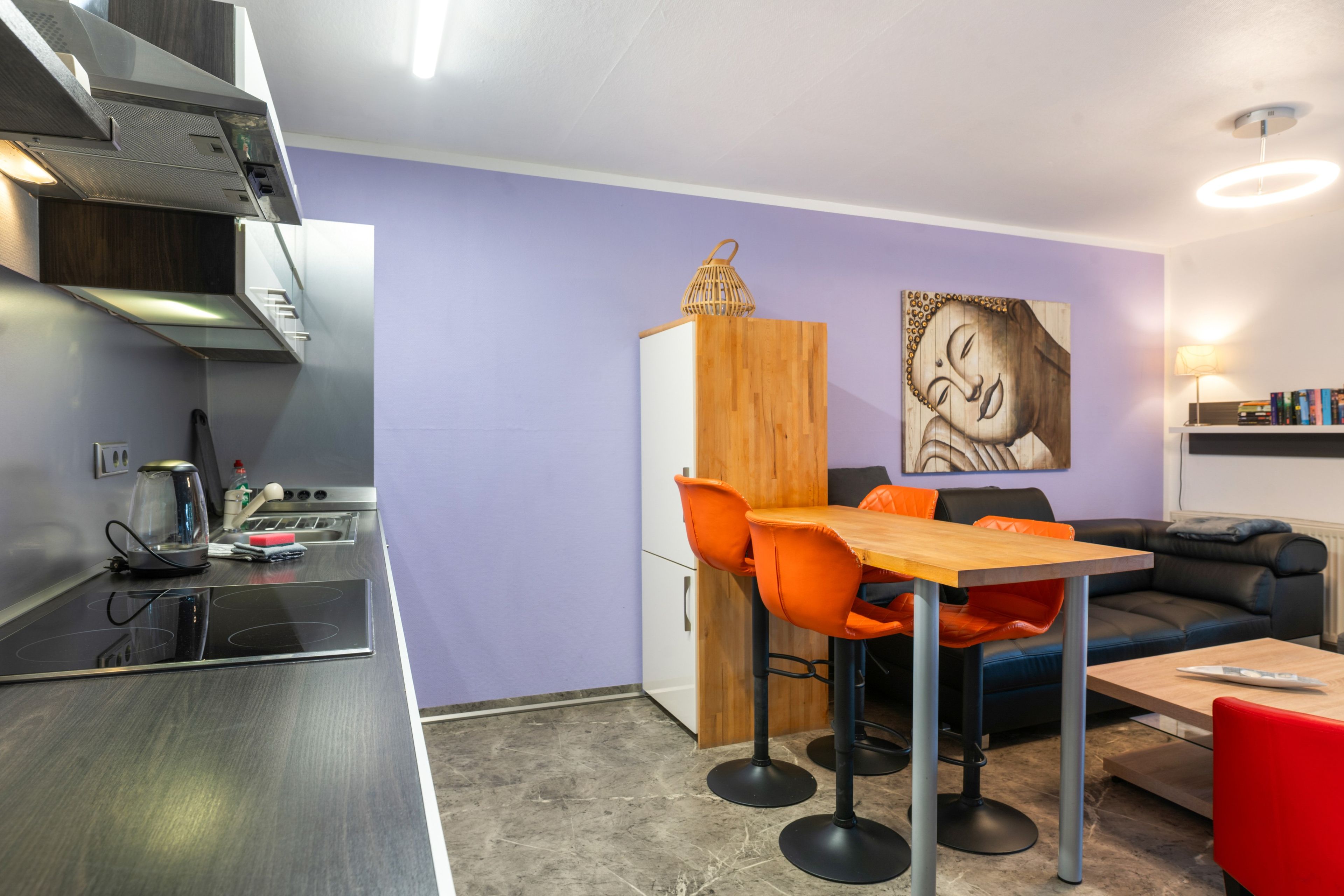 A compact kitchen with modern appliances, a dining area with a wooden table and orange chairs, and a living space with a sofa and a wall-mounted artwork.