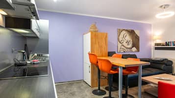 A compact kitchen with modern appliances, a dining area with a wooden table and orange chairs, and a living space with a sofa and a wall-mounted artwork.