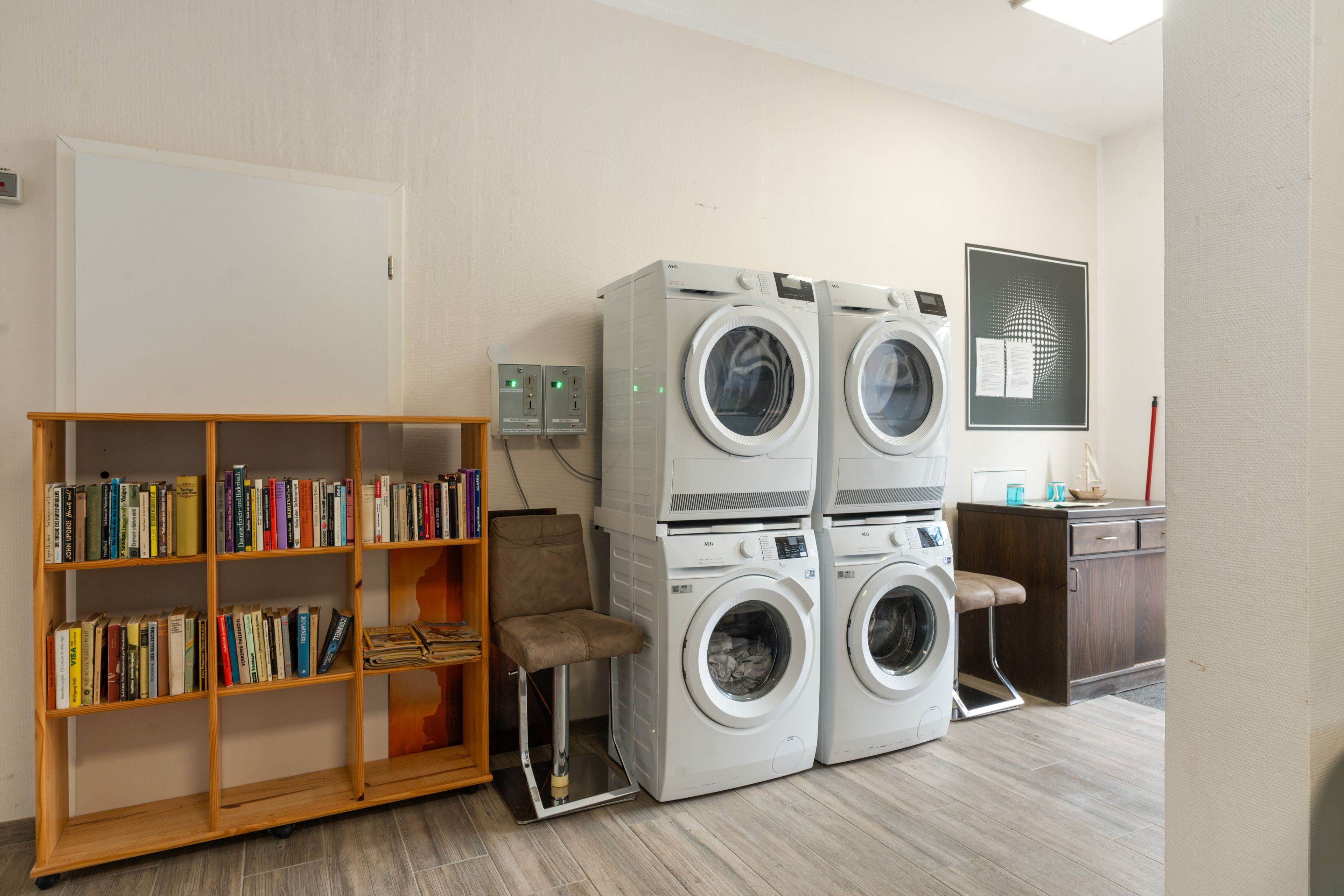 A laundry room with stacked front-loading washing machines, a wooden bookshelf with books, and a desk with a computer.