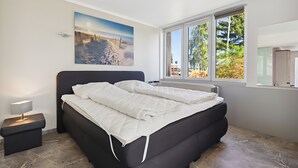 Un dormitorio con cama, una mesita de noche, una ventana con vista a árboles y un cuadro en la pared.