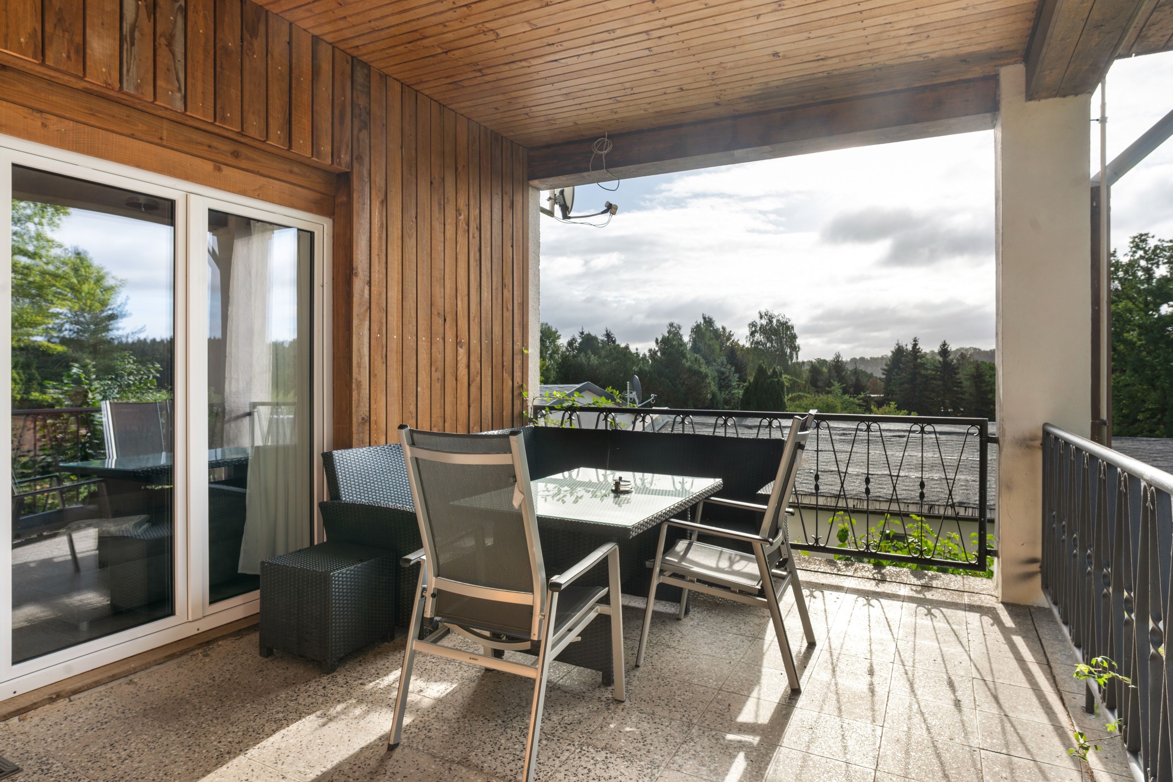 Un espace extérieur abrité, doté d’une table et de chaises, d’une porte-fenêtre coulissante et offrant une vue sur les arbres et le ciel.