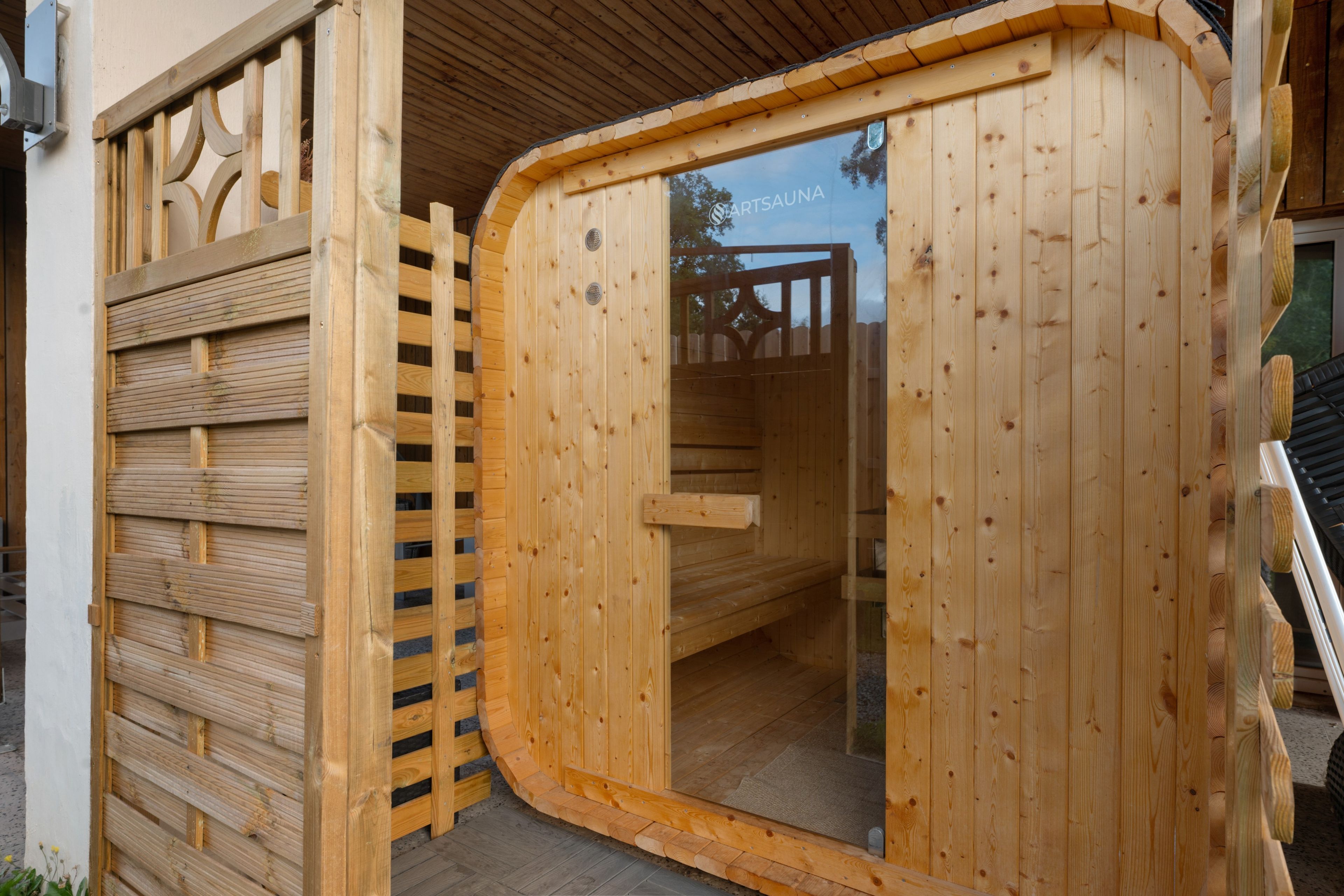 Une entrée de sauna en bois avec une porte en verre, un banc en bois à l’intérieur et une clôture en bois à l’extérieur.