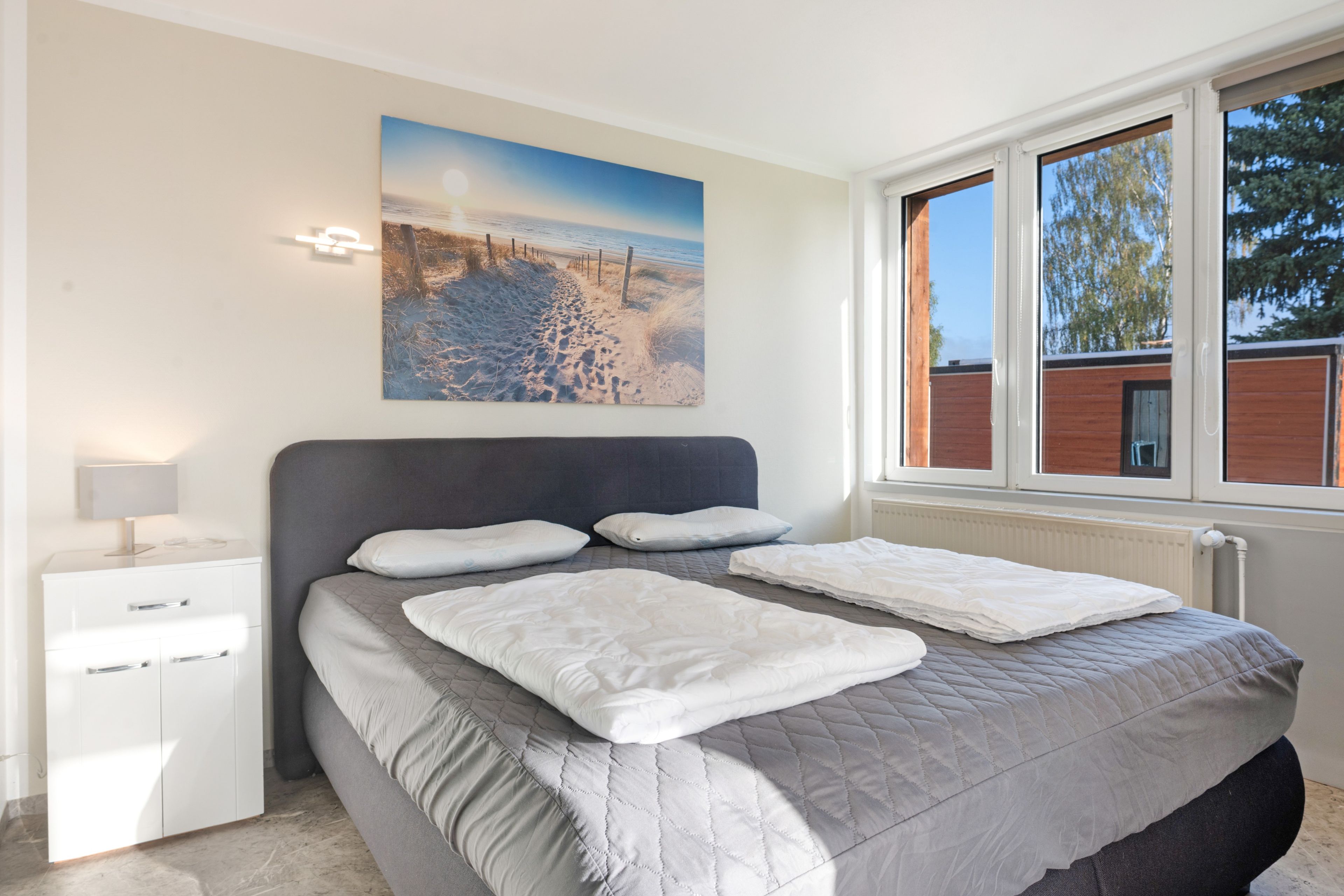 A bedroom with a bed, a nightstand, a lamp, a painting, and a window with a view of trees.