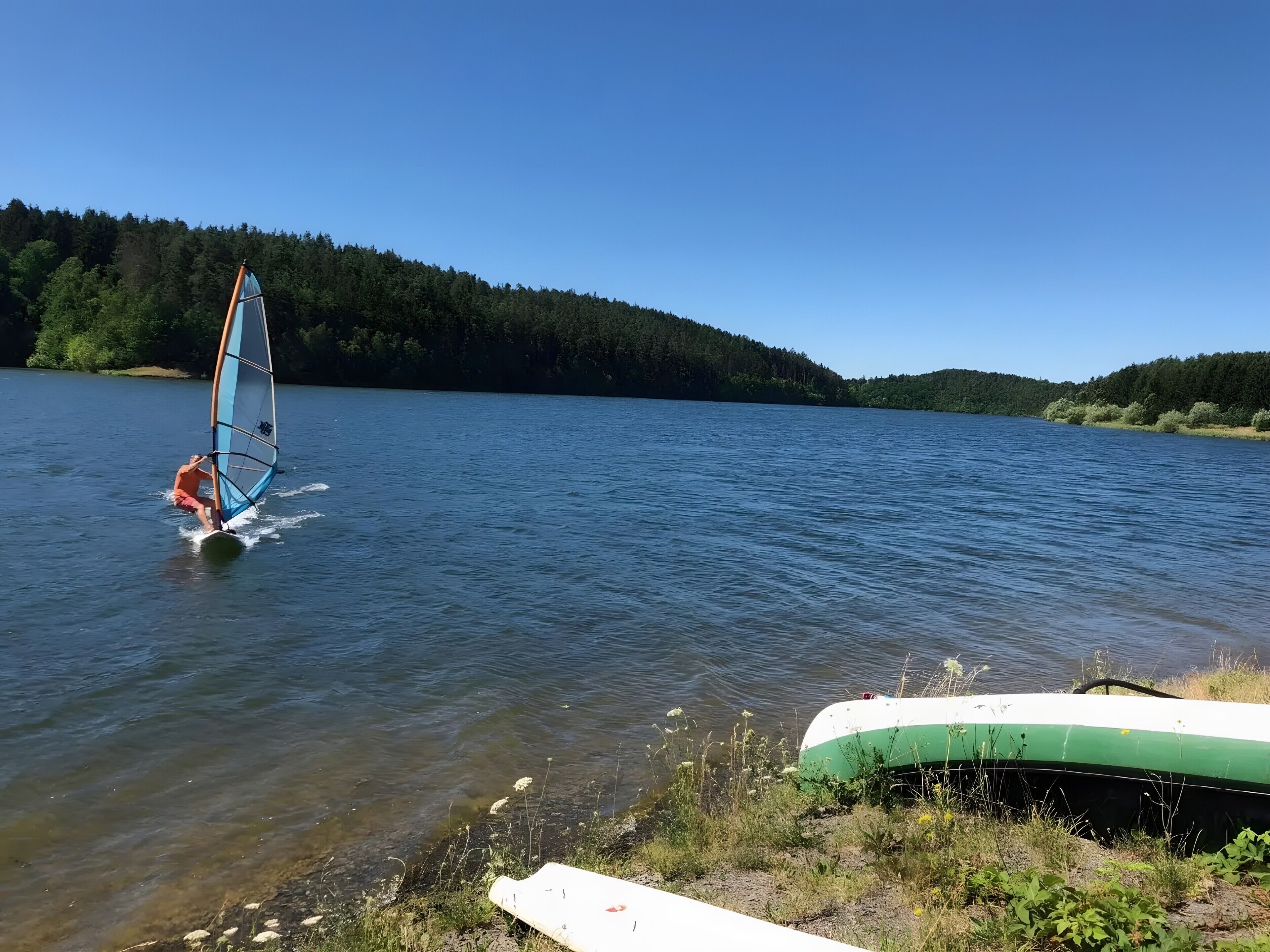 Iemand windsurft op een kalm meer met een bebost oever.