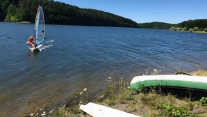 Una persona practica windsurf en un lago tranquilo con una orilla arbolada.