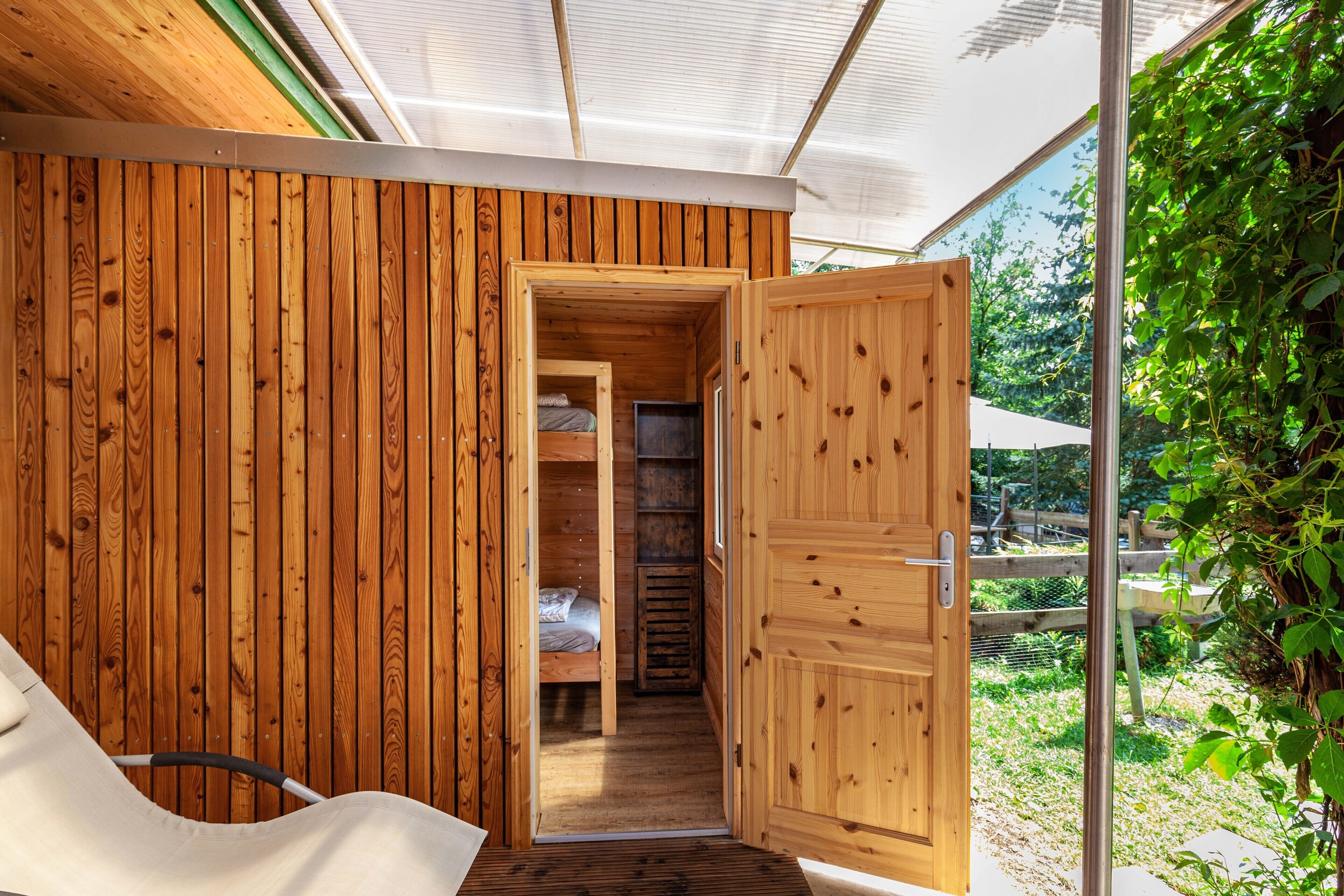 A wooden interior with a bunk bed, a door leading to an outdoor area, and a view of greenery outside.