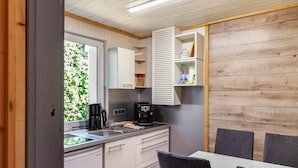 Een kleine keuken met een koffiezetapparaat, een spoelbak en kastjes. Er staat een eettafel met stoelen en een raam met beplanting erbuiten.