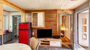 Een kleine kamer met een houten wand, een rode koelkast, een televisie, een tafeltje en een deur naar een andere ruimte.