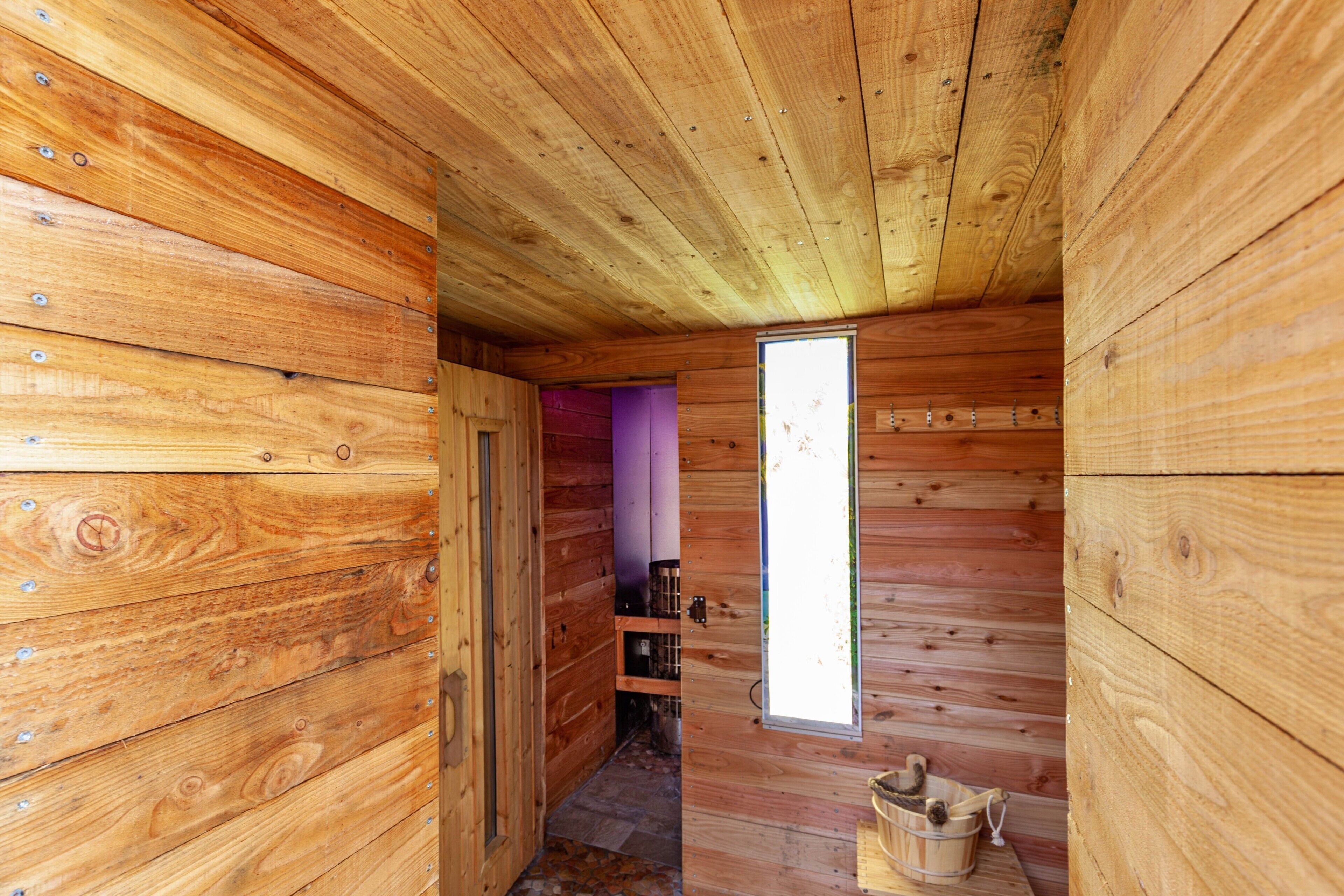 Interior de una cabaña de madera con una puerta que da a una habitación morada, una ventana y un banco de madera.