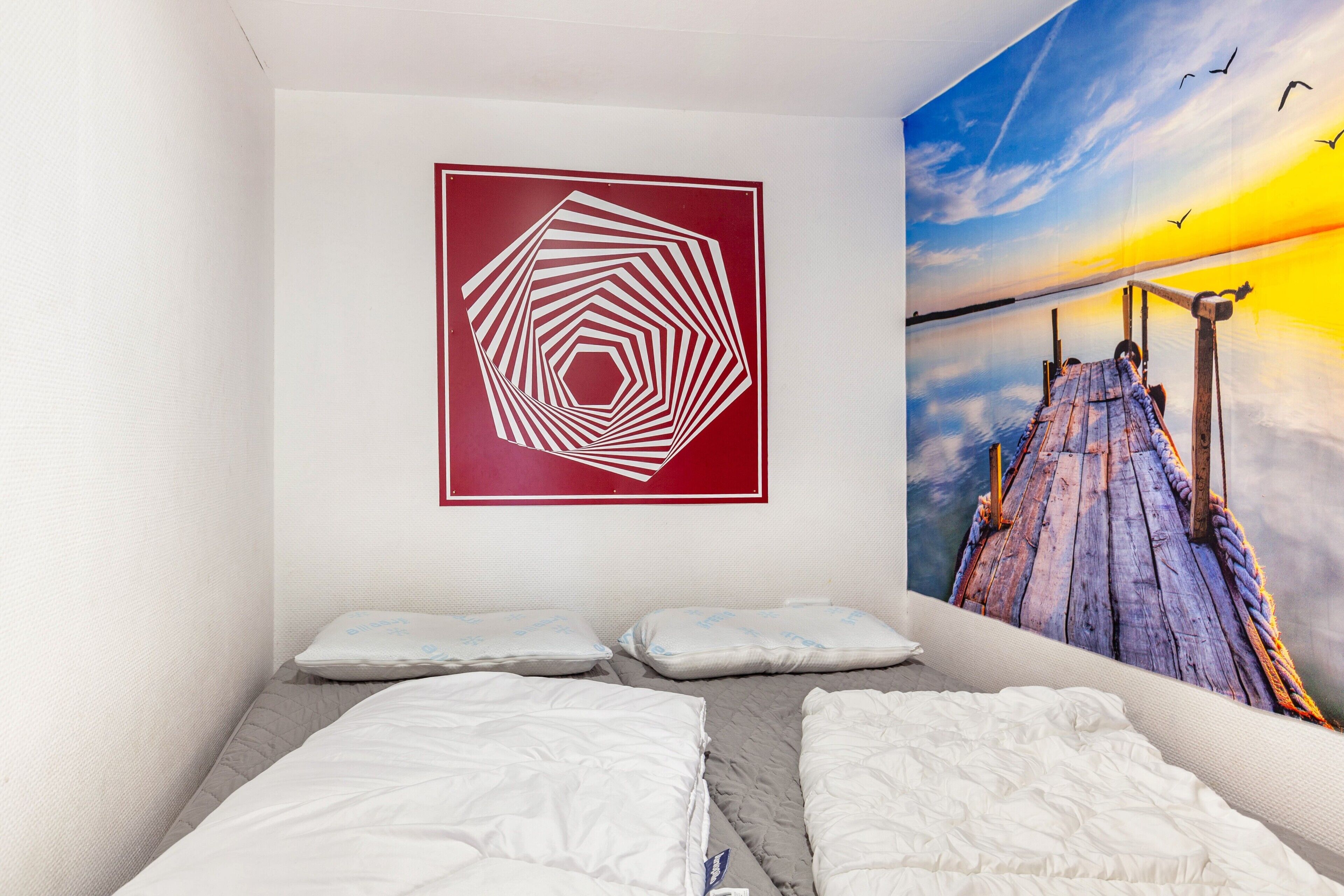 Ein Schlafzimmer mit einem Bett, Kissen, einem Wandbild und einem Wandgemälde mit Sonnenuntergang über einem Steg.
