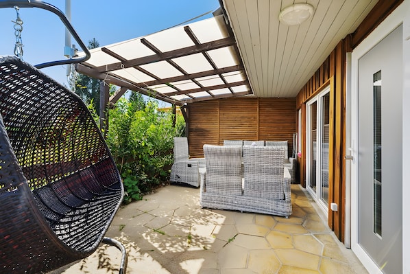 A patio with wicker furniture, a hanging chair, and a covered area with a wooden wall and glass doors.