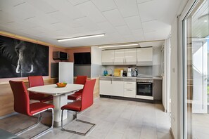 A modern kitchen with white cabinets, a dining table with red chairs, a flat-screen TV, and a large window.