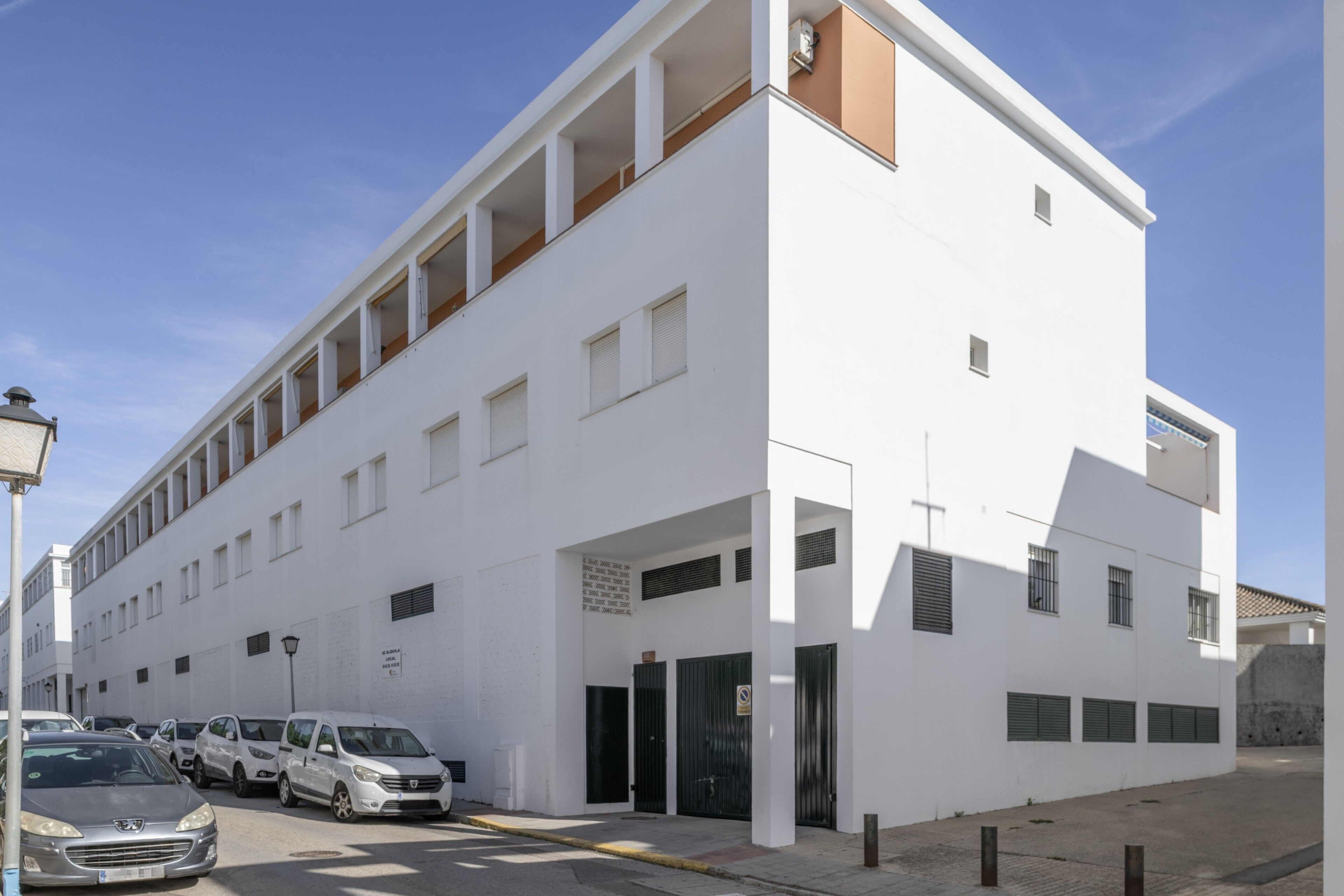 A white building with a flat roof, multiple windows, and a parking area with cars.