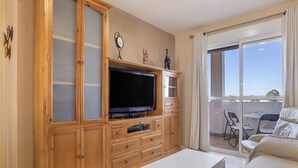 Ein Wohnzimmer mit Fernseher, Holzschränken, einem weißen Couchtisch und einem Balkon mit Sitzgelegenheiten.