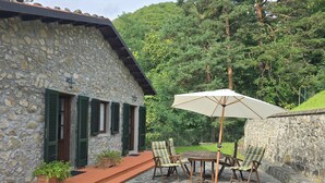 Un bâtiment en pierre avec des volets verts, une terrasse avec une table et des chaises, et une vue sur une colline boisée.