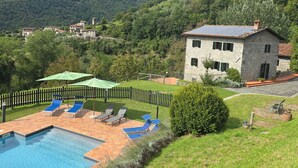 Un bâtiment en pierre doté d’une piscine, entouré d’une terrasse clôturée avec des chaises longues et un parasol vert.