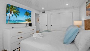 Une chambre à coucher avec un grand lit, des murs blancs, un téléviseur à écran plat, une commode et une porte.