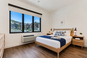Un dormitorio con una ventana grande, una cama, mesitas de noche, un radiador y vistas a edificios.