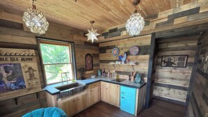 A rustic kitchen with wooden walls, a sink, and blue cabinets. There are decorative plates on the wall and a window with a view of greenery. - Luminary Lodge: an Appalachian arcade of interactive inventions above the clouds (Sevierville)