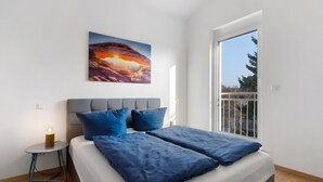 A modern bedroom with a bed, pillows, a nightstand, a painting, and a window.