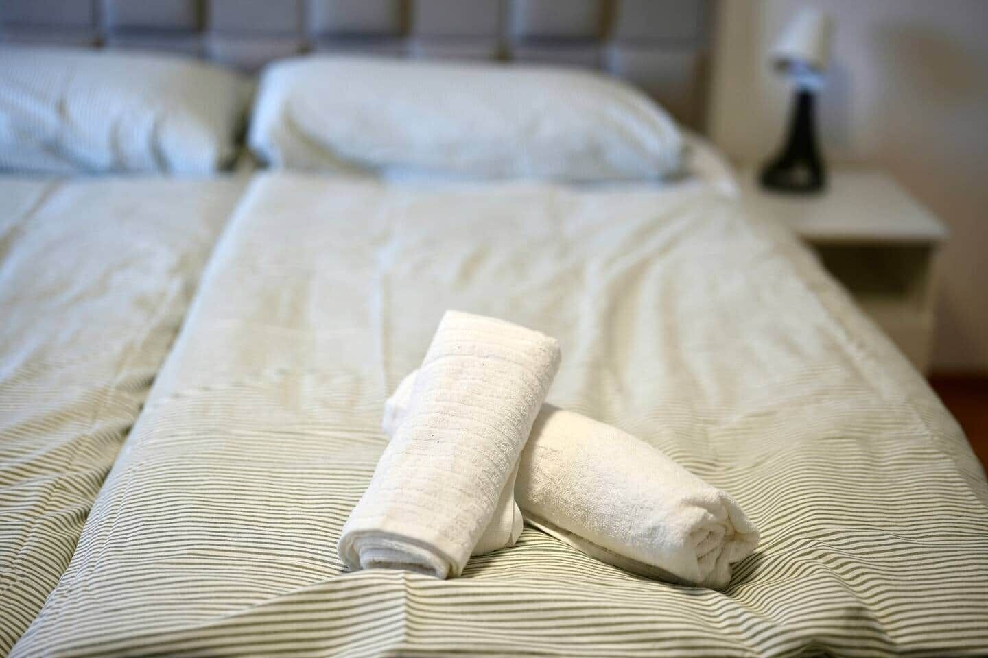 Uma cama bem arrumada com uma toalha dobrada e um travesseiro, uma mesa de cabeceira com abajur e um criado-mudo.