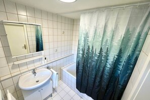 Une salle de bain avec un lavabo blanc, un miroir, une baignoire et un rideau de douche à motifs de forêt.