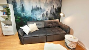 A living room with a dark leather sofa, a white side table, a bookshelf, and a forest-themed wall mural.