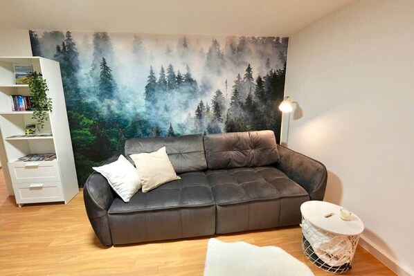 Un salon avec un canapé en cuir foncé, une table d’appoint blanche, une bibliothèque et une fresque murale sur le thème de la forêt.