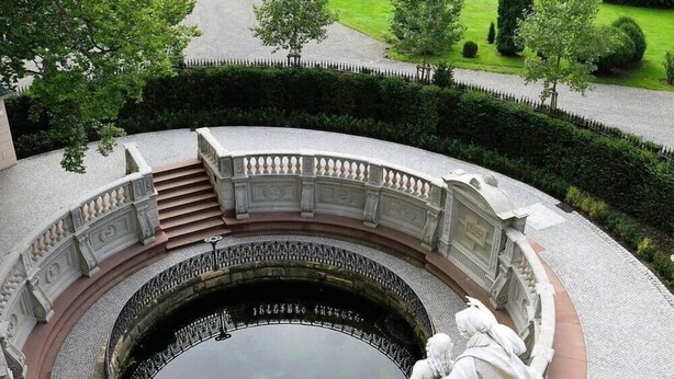 A circular fountain with statues, surrounded by a stone balustrade and a well-manicured garden.