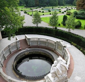 Ein runder Brunnen mit Statuen, umgeben von einer Steinbalustrade und einem gepflegten Garten.