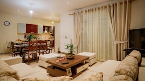A living room with a sofa, a coffee table, and a TV. There is a dining area with chairs and a table, and a kitchen in the background.