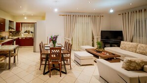 A modern living room with a dining table, sofa, TV, and kitchen in the background.