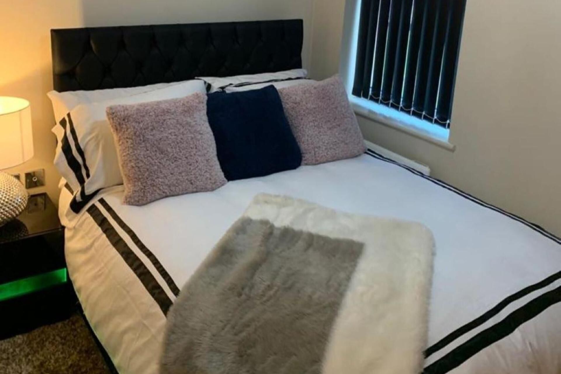 Una cama bien tendida con una manta blanca y gris, un cabecero negro y una ventana con persianas.